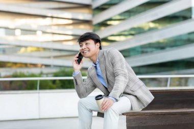 Gri ceketli ve beyaz pantolonlu Asyalı bir adam modern bir binanın dışındaki bankta oturuyor. Elinde bir fincan kahveyle telefonuyla gülümsüyor ve konuşuyor.