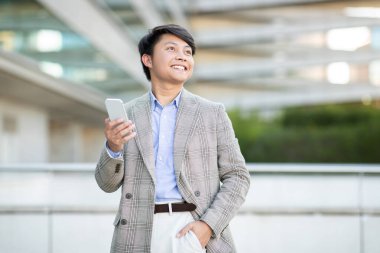 Ekose ceket ve açık mavi gömlek giymiş genç bir Asyalı şehir binasının önünde dikilirken akıllı telefonunu kullanıyor. Gülümsüyor ve fotokopi alanına bakıyor.
