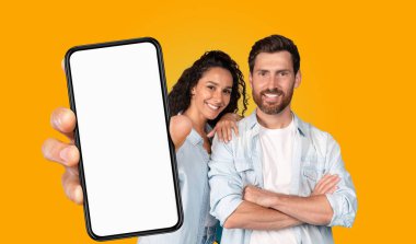 Bir çift parlak sarı bir arka planın önünde duruyor. Kadının kolu erkeğin omzunda ve ikisi de kameraya gülümsüyor. Kadın elinde boş ekranlı bir akıllı telefon tutuyor.