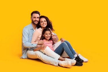 Üç kişilik bir aile, bir baba, bir anne ve kızları, parlak sarı bir arka planda birlikte oturuyorlar. Hepsi gülümsüyor ve kameraya bakıyor. Baba kolunu annesine dolamış.