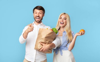 Bir adam ve bir kadın açık mavi bir arka planın önünde bakkaliye malzemelerini tutuyorlar. Kadın elinde turuncu bir poşet tutarken, adam elinde meyvelerle dolu kahverengi bir kese kağıdı tutuyor.