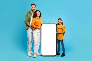 Bir baba, anne ve kızdan oluşan gülümseyen bir aile mavi bir arkaplan önünde bir arada dururlar. Ebeveynler elinde boş ekranlı büyük bir akıllı telefon tutuyorlar.