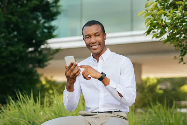 stock image A young black man, wearing a white long-sleeved shirt and khaki pants, sits outdoors in front of a modern building with lush greenery surrounding him. He is smiling and using his smartphone