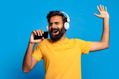 Kulaklık takan Hintli sakallı adam gülümseyerek ve bir kolunu havaya kaldırarak akıllı telefonuyla müzik eşliğinde şarkı söylüyor. Sarı bir tişört giyiyor ve mavi arka planın önünde duruyor..