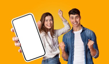 İki genç insan, bir erkek ve bir kadın, som sarı bir arka planın önünde duruyorlar. Kadın elinde boş ekranlı bir akıllı telefon tutuyor. Adam onun yanında duruyor.