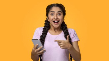 Evet, harika haberlerim var. Heyecanlı Hintli Kadın Portresi Kazananı Kutluyor, Akıllı Telefon Kullanıyor ve Gadget 'ı Gösteriyor, Gülümseyen Genç Bayan Başarılı Oluyor, Cep Telefonu Tutuyor, Sarı Stüdyo Arkaplan