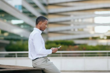 Beyaz gömlekli ve bej pantolonlu Afro-Amerikan bir adam modern bir ofis binasının dışındaki ahşap bir bankta oturuyor. Akıllı telefonunu kullanıyor, ekrana bakıyor, boşluğu kopyalıyor.