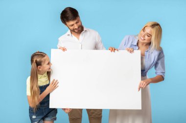 Üç kişilik bir aile, bir baba, bir anne ve kızları, açık mavi bir arkaplanın önünde büyük beyaz bir tabela tutuyorlar..