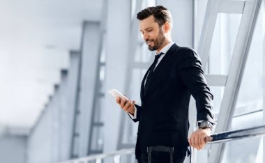 Yakışıklı sakallı iş adamı uçuş beklerken e-postayı kontrol ediyor, havaalanının penceresinin yanında duruyor, kopyalama alanı. Akıllı telefon kullanarak şık takım elbiseli orta yaşlı girişimci