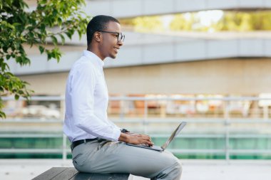 Gözlüklü ve beyaz gömlekli genç bir Afro-Amerikan adam dışarıda bir bankta oturuyor ve bir dizüstü bilgisayar üzerinde çalışıyor. Yüzünde bir gülümsemeyle yan tarafa bakıyor, boşluğu kopyalıyor.