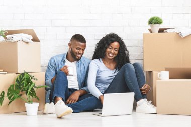 Taşınma konsepti. Mutlu siyah çift yeni daire için mobilya alıyor, yerde oturuyor ve dizüstü bilgisayar kullanıyor, boş alanı olan panorama kullanıyor.