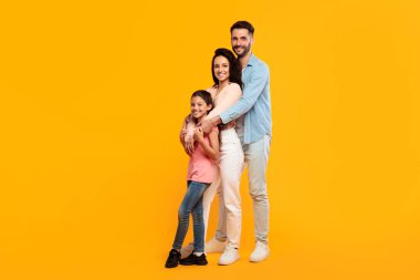 Üç kişilik bir aile, parlak sarı bir arka plana karşı birlikte poz veriyor. Baba, anne ve küçük kız gülümsüyor ve kameraya bakıyor.
