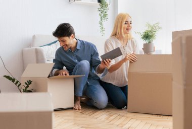 Genç bir çift diğer kutularla çevrili yeni evlerinde kutuları açıyor. Gülümsüyorlar ve yeni yerleri için heyecanlı görünüyorlar..