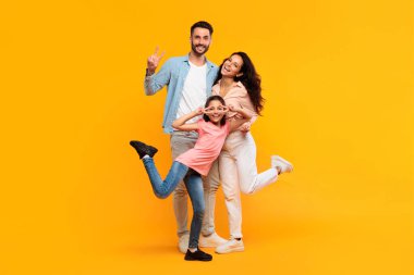 Üç kişilik mutlu bir aile sarı arkaplan önünde birlikte poz veriyor. Baba sol tarafta, eliyle barış işareti yapıyor, anne ise sağ tarafta, parlak bir gülümsemeyle..