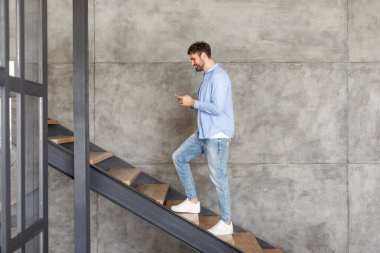 Açık mavi gömlekli ve kot pantolonlu bir adam akıllı telefonuna bakarken modern beton bir merdivende yürüyor..