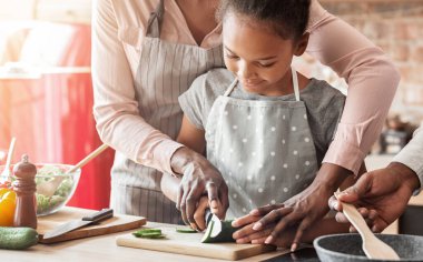 Yemek pişirmenin ilk adımları. Hevesli küçük siyah kız mutfakta annesinin gözetiminde salatalık kesiyor, kapat.