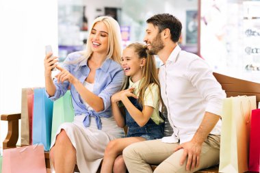 Üç kişilik mutlu bir aile alışveriş merkezindeki bir bankta oturup akıllı telefondan selfie çekiyor. Anne telefonu tutuyor, gülümsüyor ve kameraya bakıyor.