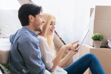 Yeni bir yere taşındıktan sonra taşınan kutuların arasında oturan neşeli çift tablet bilgisayar kullanıyor. Boş Boşluk