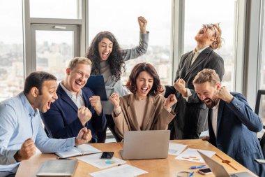 Çeşitli iş adamlarından oluşan bir ekip, şehrin ufuk çizgisine bakan geniş pencereli modern ofis ortamında başarıyı kutluyor. Hepsi gülüyor ve tezahürat yapıyor, yumrukları havada.