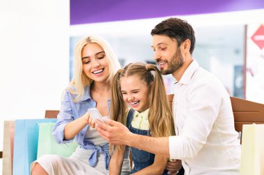 Üç kişilik bir aile bir alışveriş merkezinde oturmuş akıllı telefona bakıyor. Ebeveynler gülümsüyor ve küçük kız gülüyor. Etrafları alışveriş torbalarıyla çevrili..