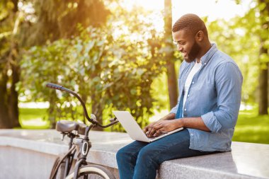 Bedava Wifi Spot. Dışarıda dizüstü bilgisayarda çalışan genç Afrikalı Amerikalı bir adam park yerinde, yanında motosikletiyle oturuyor.