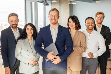 A group of smiling business professionals stand together in an office setting. The man in the center of the group is holding a laptop, while the others stand around him with their arms crossed clipart