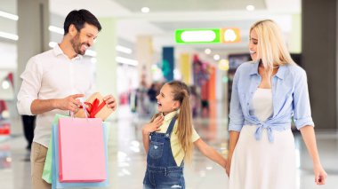 Elinde alışveriş poşetleri olan bir baba ve kırmızı kurdeleli bir hediye heyecanlı kızına bunu gösteriyor, karısı onların yanında yürürken. Üçü modern bir alışveriş merkezinde alışveriş yapıyorlar.