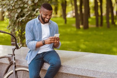 Neşe dolu Afrikalı Amerikalı bisikletini bankın yanına park etmiş ve parkta akıllı telefon kullanıyor. İnternete mesaj atıyor ya da internette geziniyor.