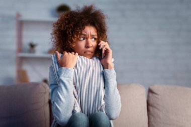 Korkmuş afro kadın telefon ediyor ve kanepede yastığı kucaklıyor, evin yakınındaki gürültüden korkuyor, boş yer var.