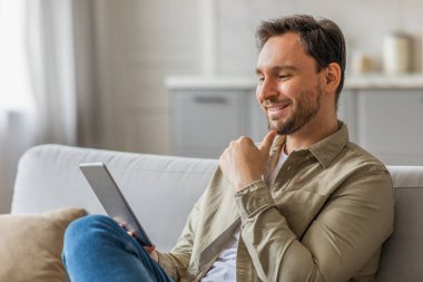 Oturma odasındaki beyaz bir kanepede tablet kullanan bir adam oturuyor. Tişört ve kot pantolon giymiş. Yüzünde rahat bir ifade var. Uzayı taklit ediyor.