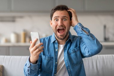 Mavi düğmeli gömlekli bir adam, sol elinde bir akıllı telefonu tutarken ve sağ elini kafasına dayarken bir koltukta şaşkın şaşkın oturuyor. Ağzı açık kaldı..