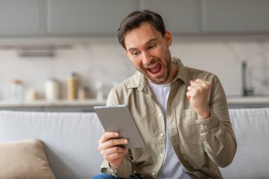 Kanepede oturan bir adam elinde tabletle heyecanla tepki veriyor. Ağzı kocaman bir gülümsemeyle açık ve yumruğunu havaya kaldırıyor. Bir zaferi kutluyor gibi görünüyor.