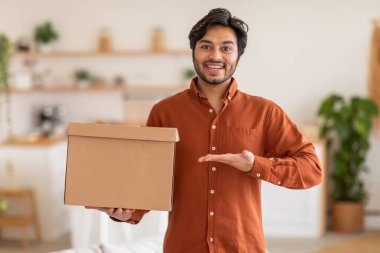 Arap adam önünde kocaman bir gülümsemeyle bir karton kutu tutuyor ve avuç içi kameraya doğru açık. Arka plan bulanık ve bulanık. Bu da bir ev ya da ofis ortamı olduğunu gösteriyor..