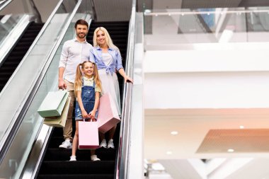 Mutlu bir aile, bir baba, bir anne ve küçük bir kız modern bir alışveriş merkezinde yürüyen merdivene biniyorlar. Hepsi gülümsüyor ve ellerinde alışveriş torbaları, kopyalama alanı