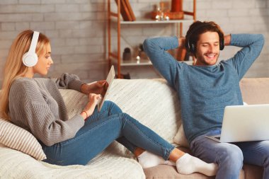 Laptop ve tablet bilgisayar kullanan rahat bir çift içeride koltukta oturuyor. Hafta Sonu Evde.