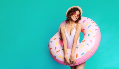 Gülümseyen siyah bir kadın, önünde pembe donut şeklinde şişme bir havuz tutan bir deniz mavisi zemininin önünde duruyor. Beyaz bir mayo, güneş gözlüğü ve hasır şapka giyiyor..
