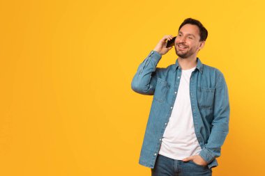 Kot gömleğin altına beyaz t-shirt giyen bir adam, parlak sarı bir arka planın önünde duruyor, akıllı telefonuyla gülümserken konuşuyor. Cebinde bir eli var ve fotokopi odasına bakıyor.