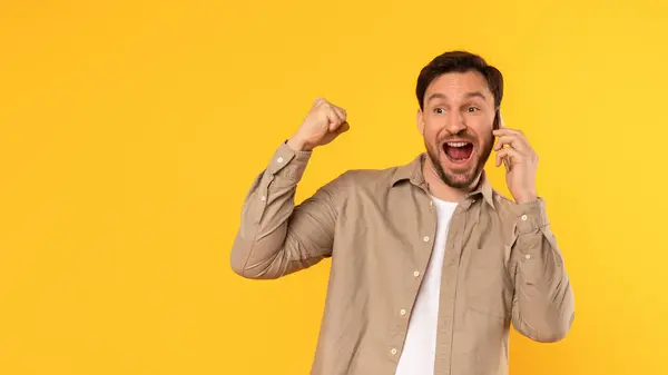 stock image A man with a beard is talking on his phone with an excited expression. He is standing in front of a yellow background and has raised his right arm in the air, copy space