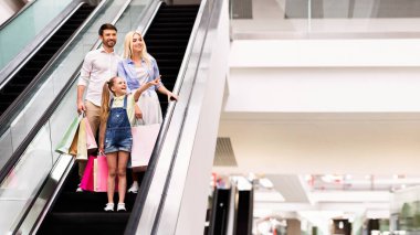 Genç bir kız, renkli alışveriş torbaları taşıyan modern bir alışveriş merkezinde ailesiyle birlikte yürüyen merdivene binerken heyecanlı bir şekilde işaret ediyor. Başarılı bir alışveriş günü, fotokopi alanı...