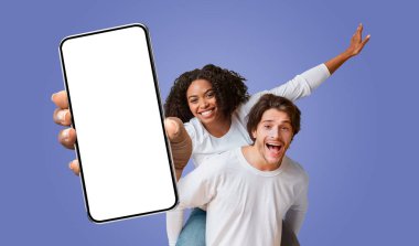 Genç, çok ırklı bir çift önlerinde boş bir ekranla akıllı bir telefon tutuyor. Kadın, erkeğin sırtındayken başparmağını kaldırıyor..