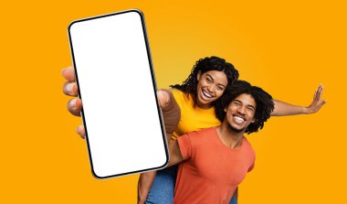 Sarı tişörtlü genç siyah bir kadın, kırmızı gömlekli bir adama sırtına biniyor. İkisi de sırıtıyor. Kadın büyük bir akıllı telefonun boş ekranını işaret ediyor.