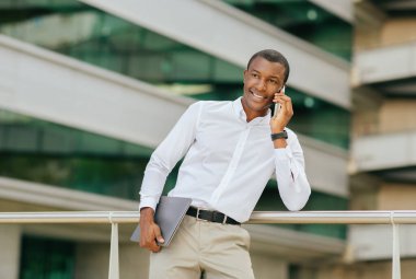 Beyaz gömlek ve haki pantolon giyen siyah işadamı modern bir ofis binasının önünde duruyor ve cep telefonuyla konuşuyor. Kolunun altında dizüstü bilgisayar tutuyor ve gülümsüyor.