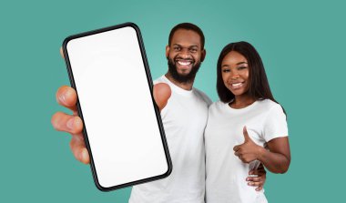 Genç bir siyah çift, deniz mavisi arkaplanının önünde duruyor. Adam elinde beyaz bir ekranla akıllı bir telefon tutuyor ve kadın başparmağını kaldırıyor, gülümsüyor..