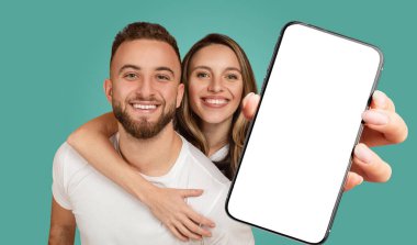 Genç bir çift turkuaz bir arka planın önünde mutlu bir şekilde gülümsüyor. Kadın kolunu erkeğin omuzlarına dolamış ve elinde boş bir ekranla akıllı bir telefon tutuyor.