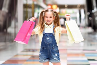 Uzun sarı saçlı genç bir kız elinde iki alışveriş torbası tutarken mutlu bir şekilde gülümsüyor, biri pembe diğeri sarı, parlak ışıklı bir alışveriş merkezinde..