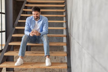 Bir adam binanın içindeki ahşap bir merdivende oturuyor, gülümsüyor ve akıllı telefonuna bakıyor. O, mavi kot pantolon, beyaz tişört ve mavi düğmeli gömlek giyiyor..