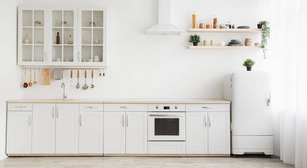 Световой скандал в кухонном интерьере. Белая мебель с посудой, полки с посудой и растения в горшках, небольшой холодильник у окна, панорама, пустое пространство