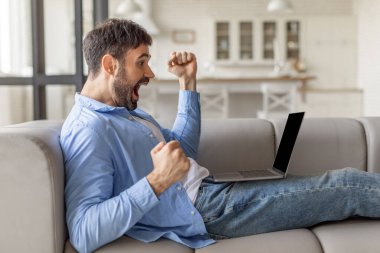 Bir adam gri bir kanepeye oturur, vücudu sola döner ve heyecanını ifade eder. Mavi düğmeli bir gömlek ve kot pantolon giyiyor ve kucağında bir dizüstü bilgisayar var..