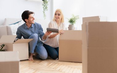 Genç bir çift yeni evlerinin zemininde birlikte kutuları paketliyor..