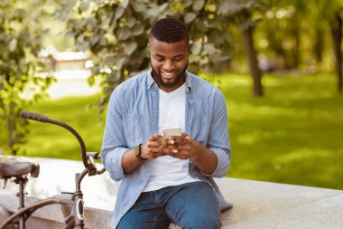 Happy african american guy in casual clothes texting on smartphone near bicycle, relaxing on bench in city park, free space